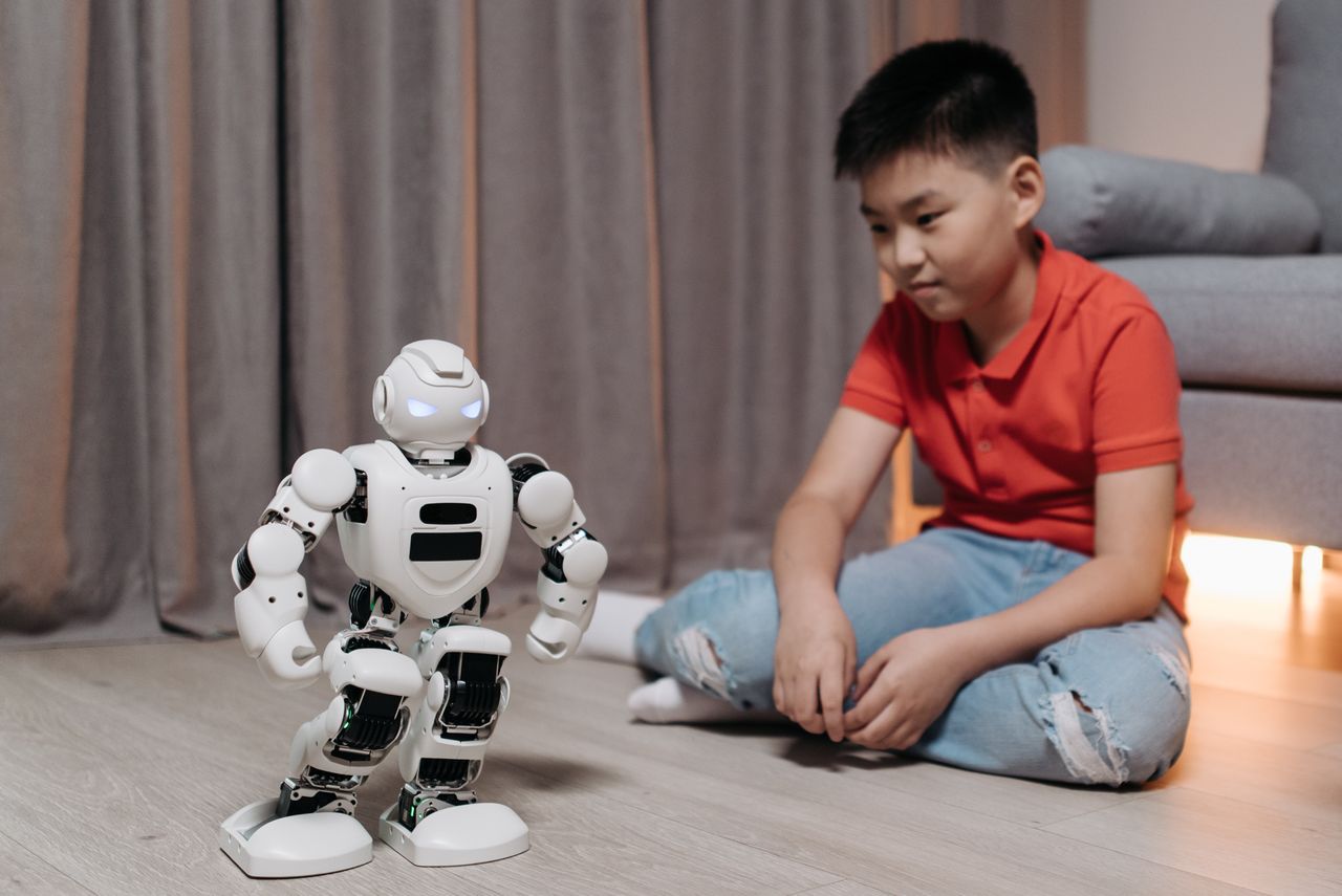 about-bg An asian boy sitting on the floor, interacting with a white robot, showcasing innovation and technology.