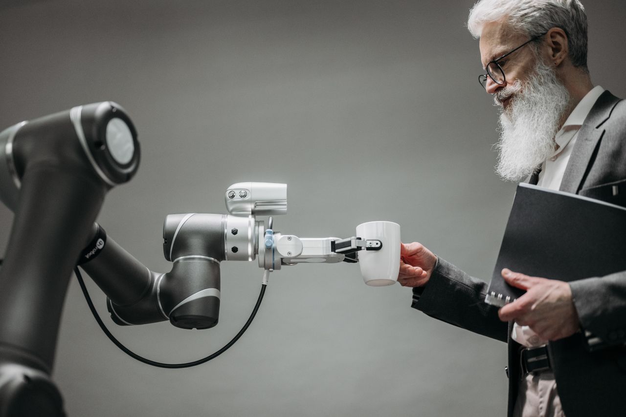 services-03 An elderly man receives a cup from a robotic arm in a modern office setting.